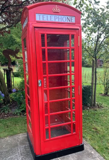 A red British telephone booth situated on a stone path with grass and trees in the background.