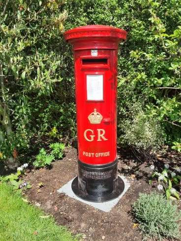 A red cylindrical postbox with "GR" and a crown emblem, set on a concrete base, surrounded by greenery and plants.