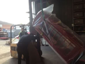 A forklift is moving a large, wrapped red object inside a warehouse, with two people assisting.
