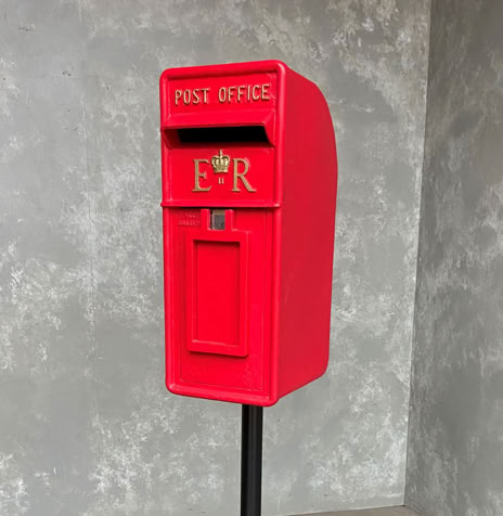 A red mail box on a black pole, set against a gray concrete wall.