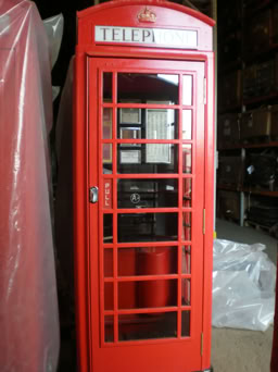 A red telephone kiosk door installed in an indoor setting with sunlight casting shadows on the wall.