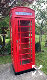 A red British telephone booth situated outdoors on a concrete surface, surrounded by grass and trees.