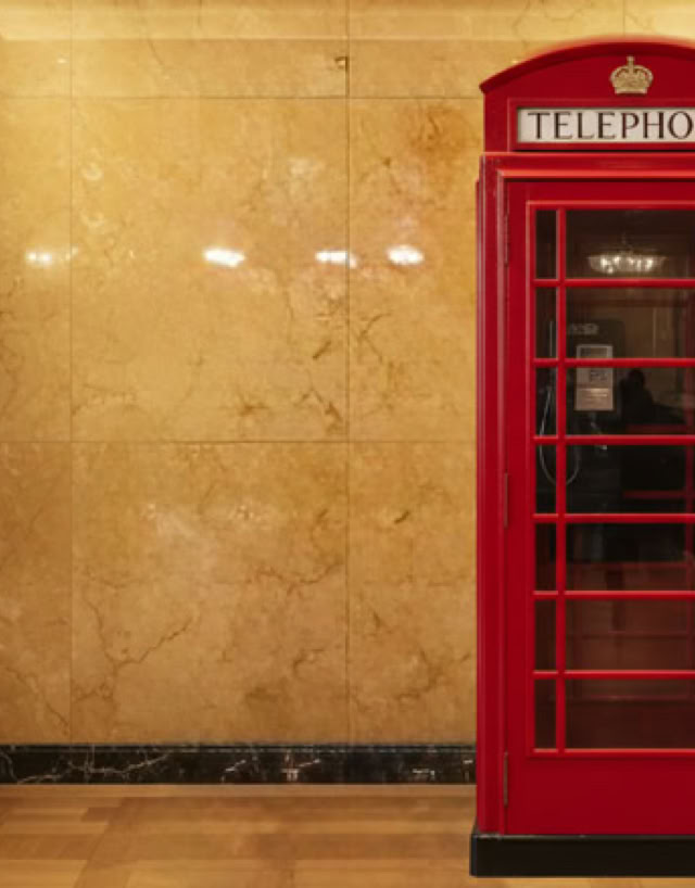 A red telephone booth against a marble wall in a hallway with wooden floors and wall sconces.