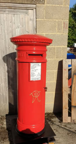 A red cylindrical post box with a slot and a label, situated against a brick wall and a wooden door.